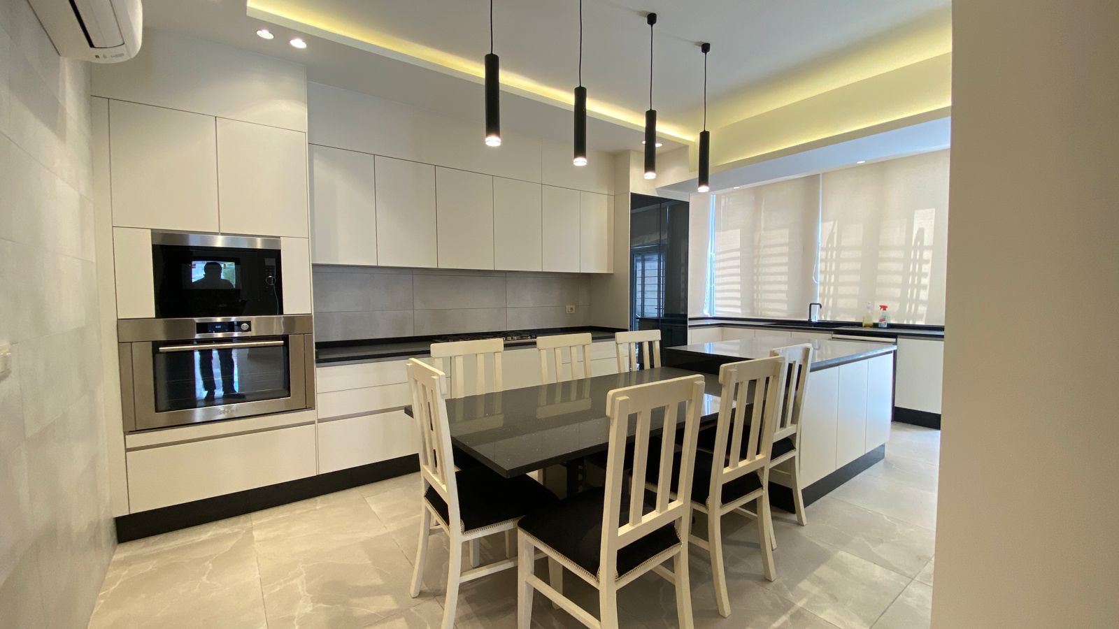 f69e704b-20ed-448e-b7ce-94ed60b1d62a Modern white kitchen with built-in stainless steel oven, black countertop island, and six white chairs around a dark table for dining.