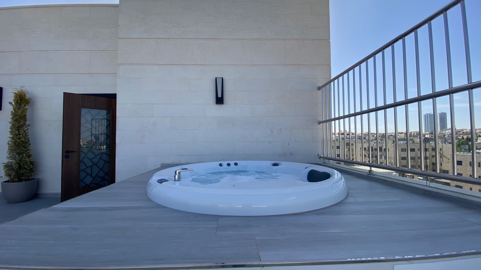 f6f08c12-4388-4b2a-b0f4-6e91264184f3 Rooftop terrace with a circular white jacuzzi on a gray wooden deck and city skyline in the distance, railing on the right.