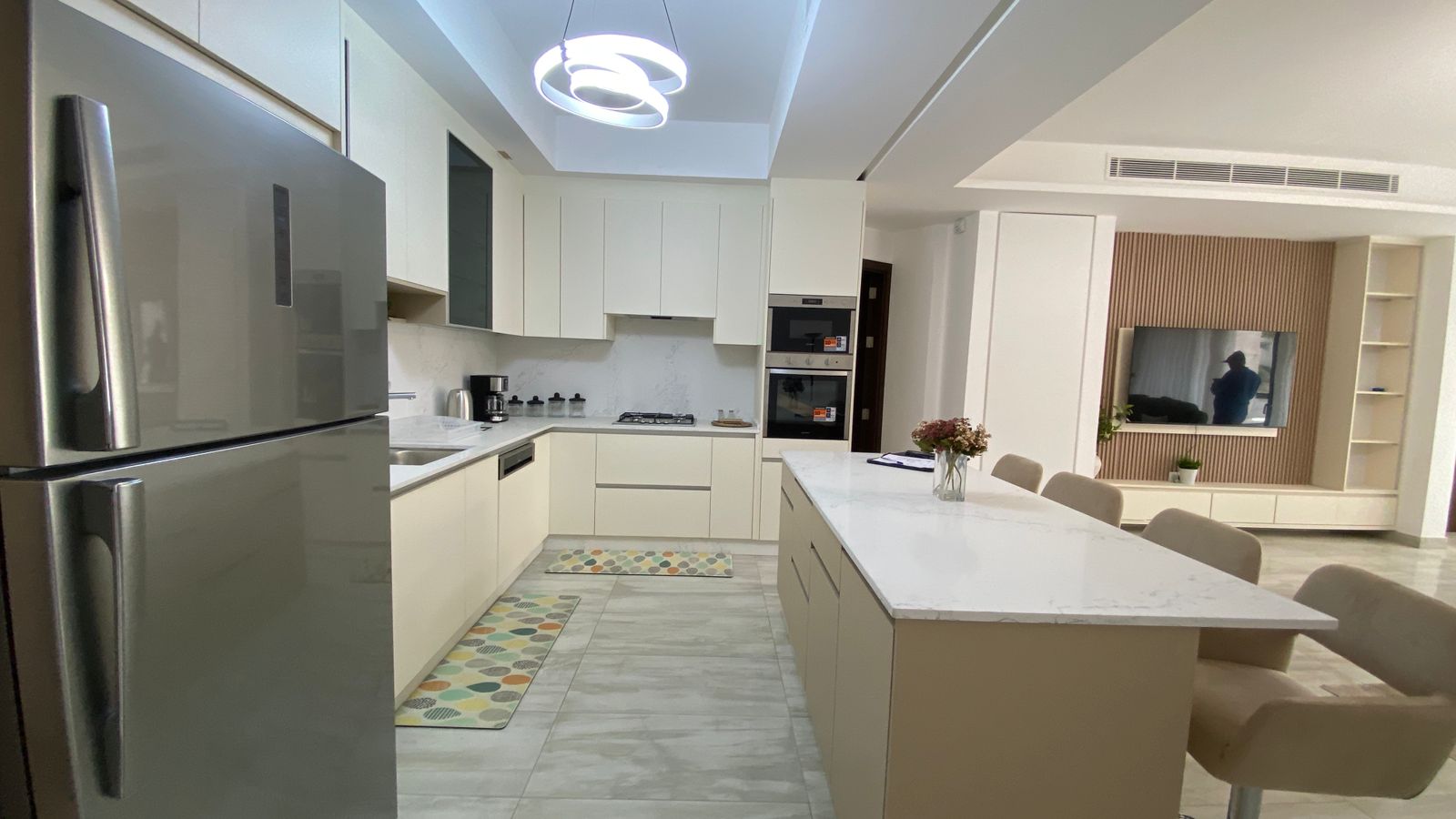 f7461a3b-dbc5-42b3-a50d-bf1ecbe17426 Open-concept kitchen with a stainless steel refrigerator, white cabinets, marble countertops, and a long island with seating; living area with a wall-mounted TV in the background.