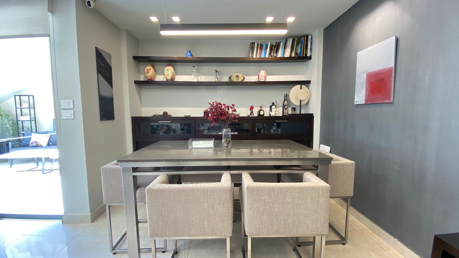 f78eb76a-923f-49ff-b9ed-200ee46ab6c6 Modern dining area with a rectangular stone-top table, four beige upholstered chairs, and a dark sideboard beneath open shelves with books and decor.