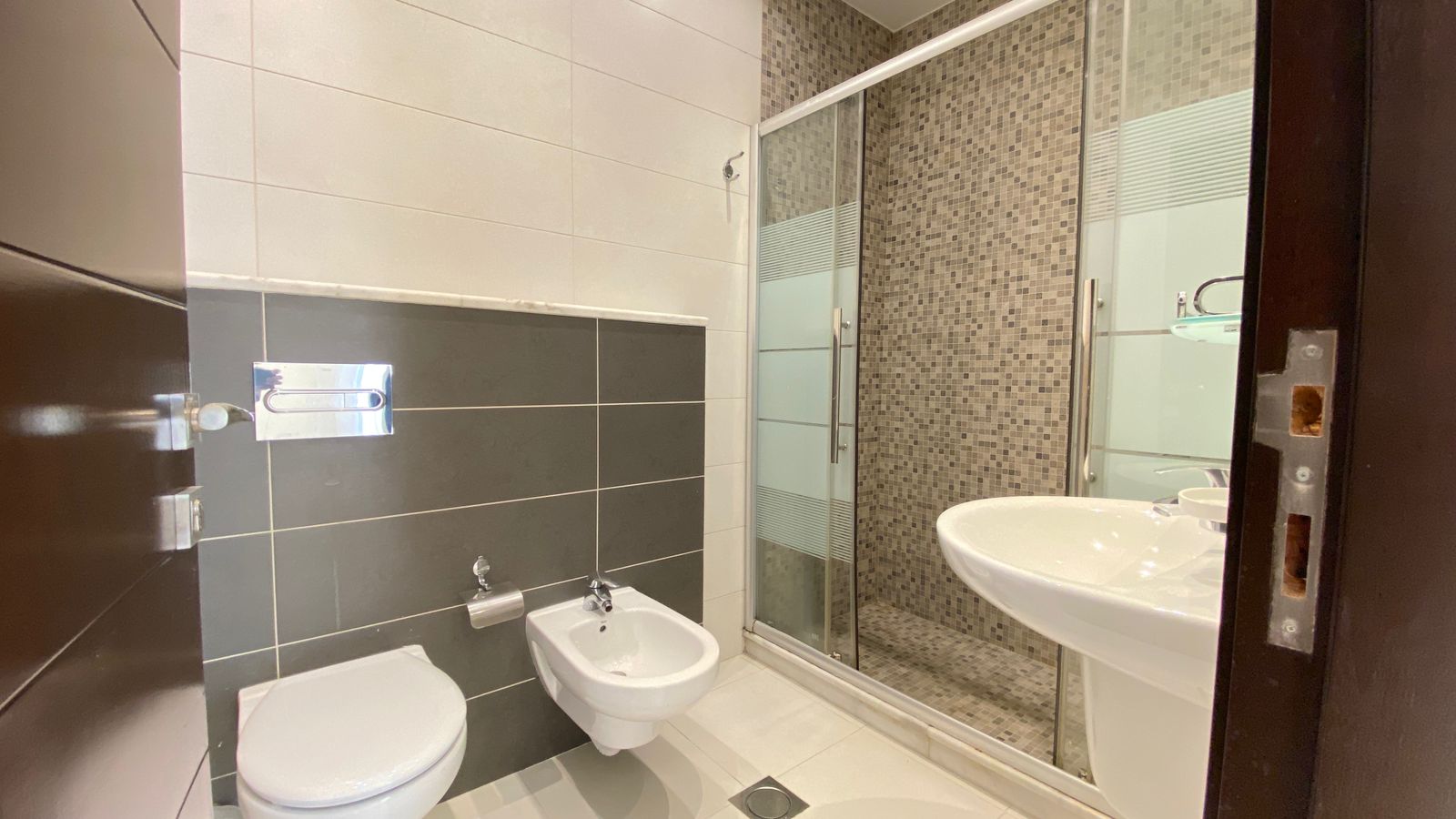 f826adad-6611-49c1-a407-975219048eca Modern bathroom featuring a white toilet and bidet opposite a glass-walled shower with mosaic tile walls; pedestal sink in front, dark gray wall tiles, and light floor tiles.