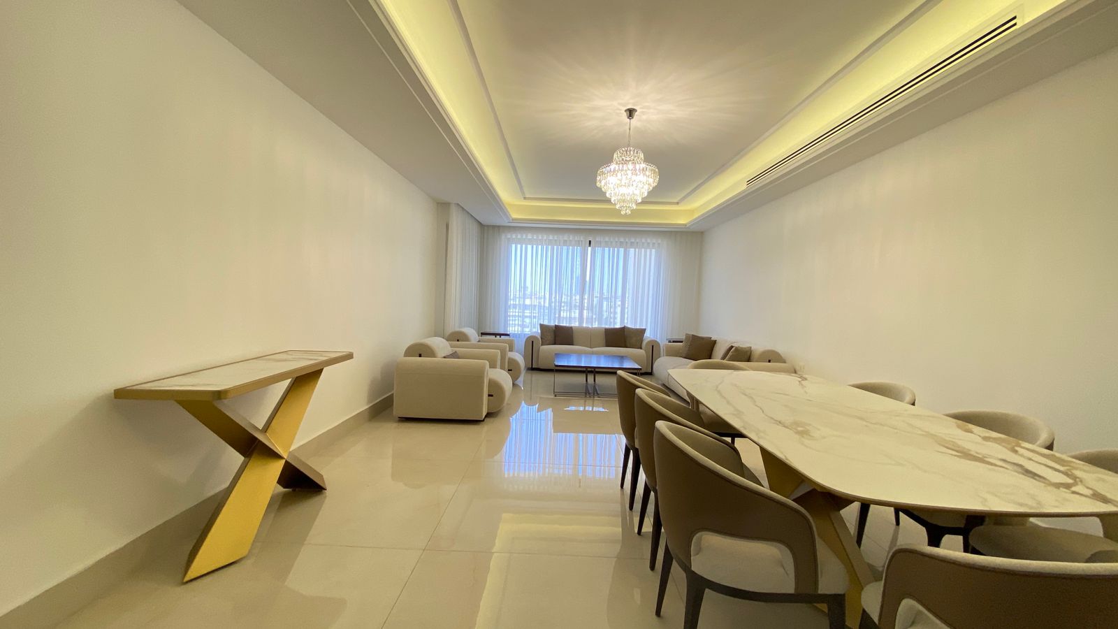f8978814-135f-4a43-a7f6-448b36c4c2a7 Bright living and dining room with a long marble table and beige chairs, cream sofa seating, and a chandelier centered in a tray ceiling.
