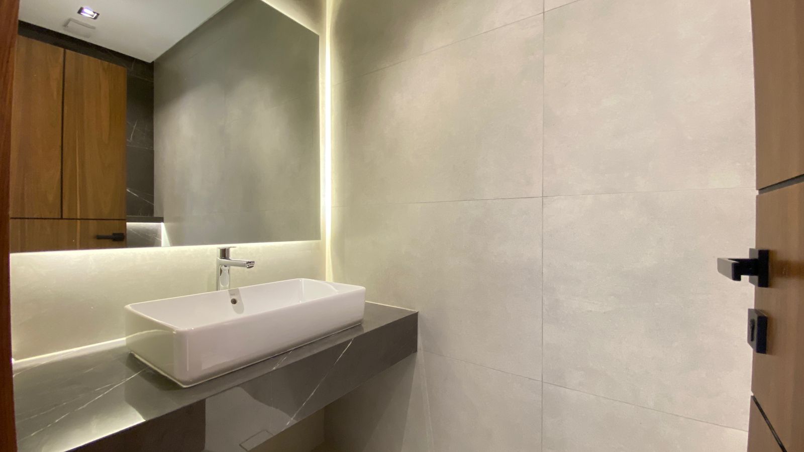 f91fc7d1-f148-4b98-91bf-6d1def3bbcf8 1 Modern bathroom vanity with a white rectangular sink on a dark countertop, beige tiled walls, and a large mirror.