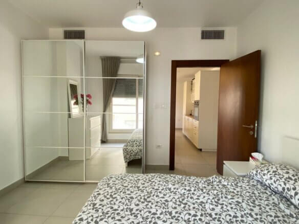 Bright modern bedroom with a large mirrored wardrobe, white chest of drawers, and a bed with grey floral bedding.