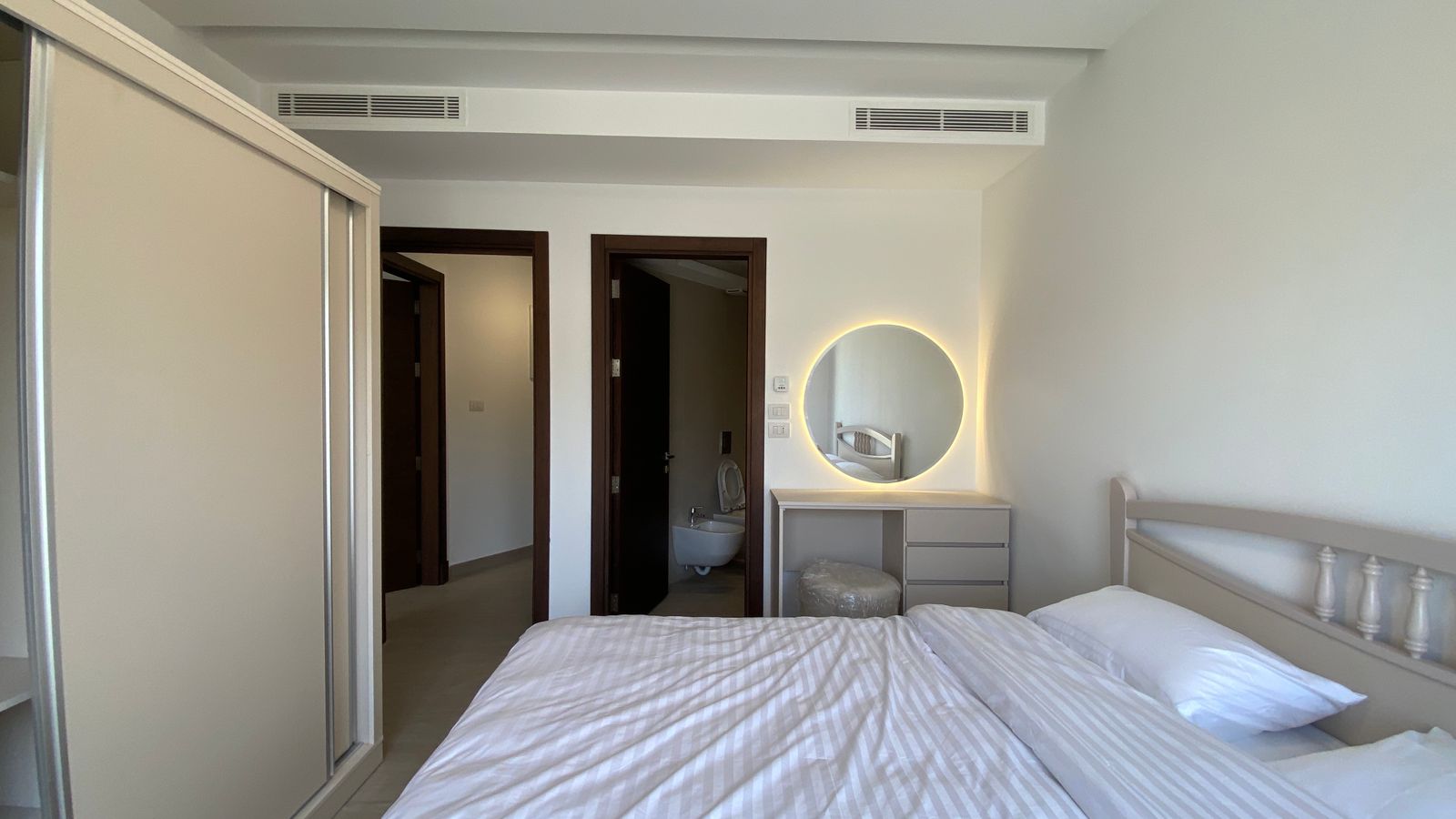 f994bdf7-57fa-4718-a81e-cf4408687579 Bright hotel-style bedroom with a white bed in foreground, a large cream wardrobe on the left, and a vanity with a round backlit mirror on the wall opposite the bed, plus a doorway leading to a bathroom where a toilet is visible.
