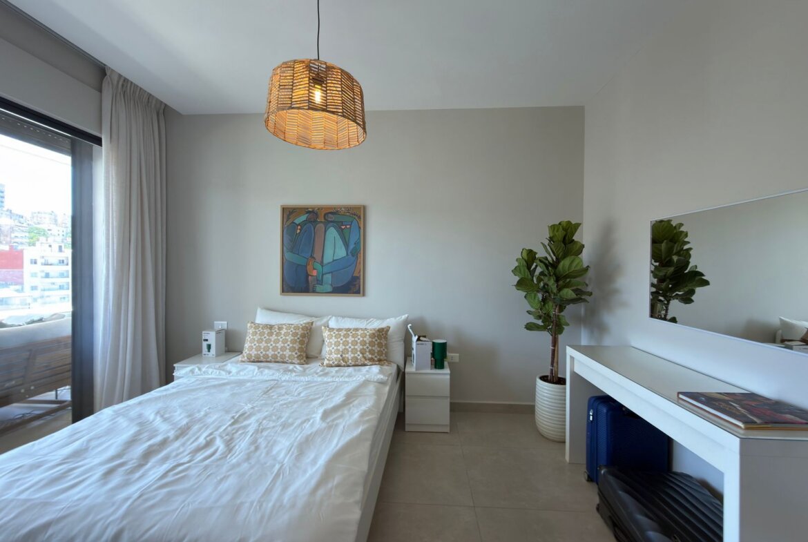 Bright, minimalist bedroom with a white bed and two beige patterned pillows, a framed abstract painting, and a woven pendant light above.