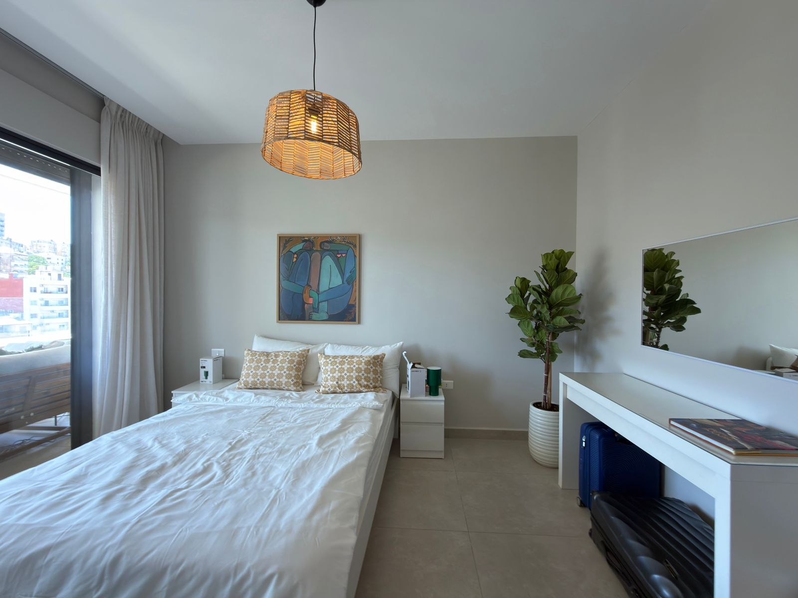f9cebddf-e7e9-4471-9571-f635d91e662f Bright, minimalist bedroom with a white bed and two beige patterned pillows, a framed abstract painting, and a woven pendant light above.