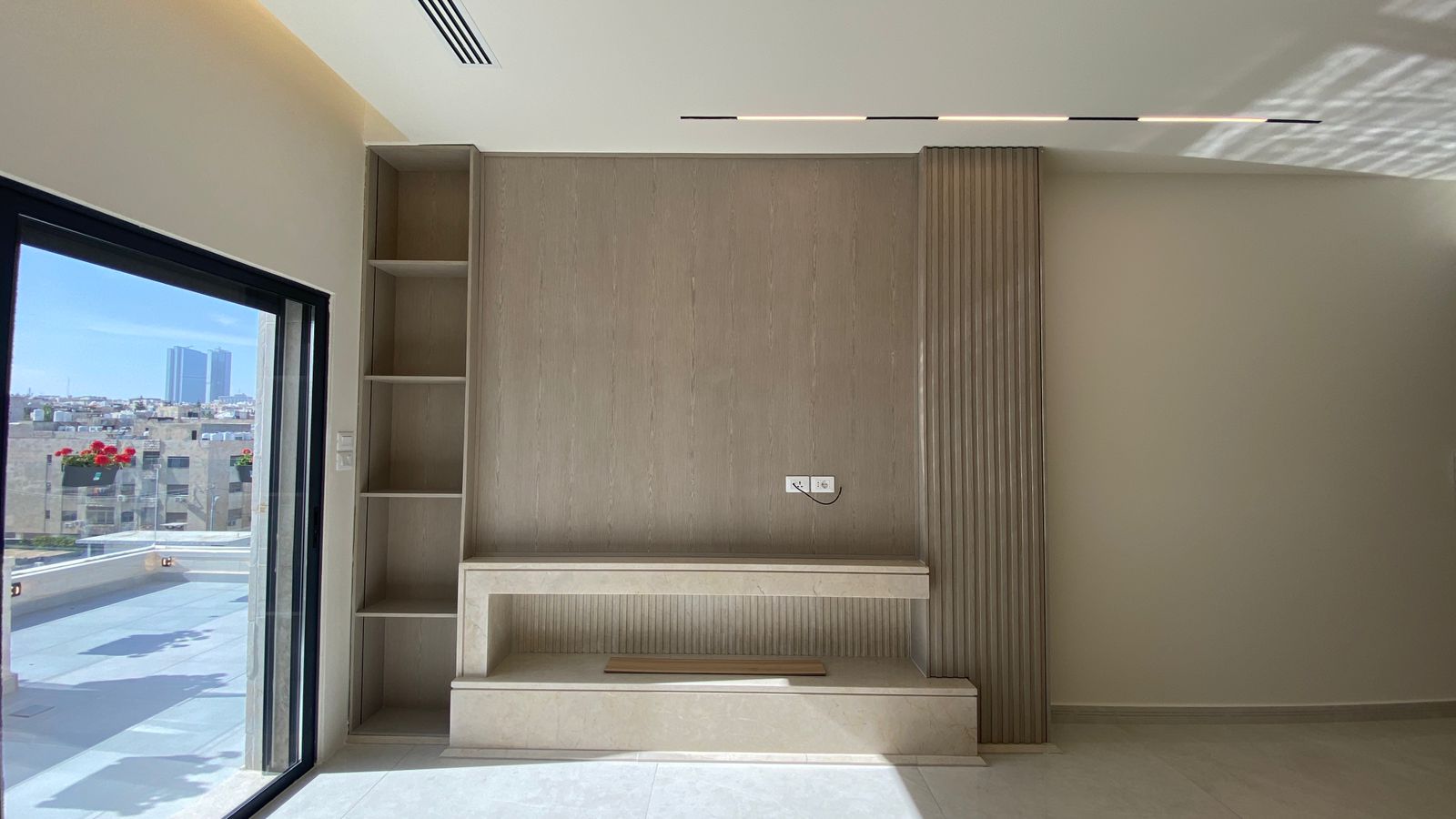 f9dad051-9936-430f-a01a-07cde94a8312 Modern living room wall unit with built-in shelves on the left, a central beige marble fireplace/bench, and vertical paneling on the right, with a large sliding glass door showing a city view outside and potted flowers on the balcony.