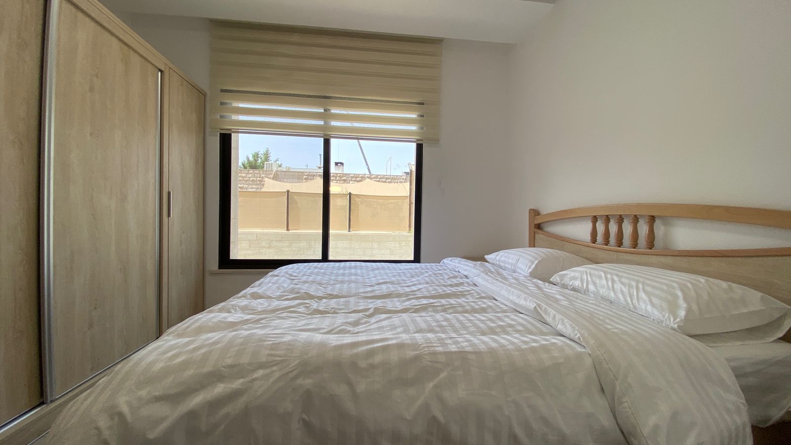 fa168cd4-e3e1-41a0-a7f7-aad47647efc2 Bedroom scene with a wooden bed frame and white striped bedding, a tall wooden wardrobe on the left, and a window with beige blinds showing an outdoor courtyard.