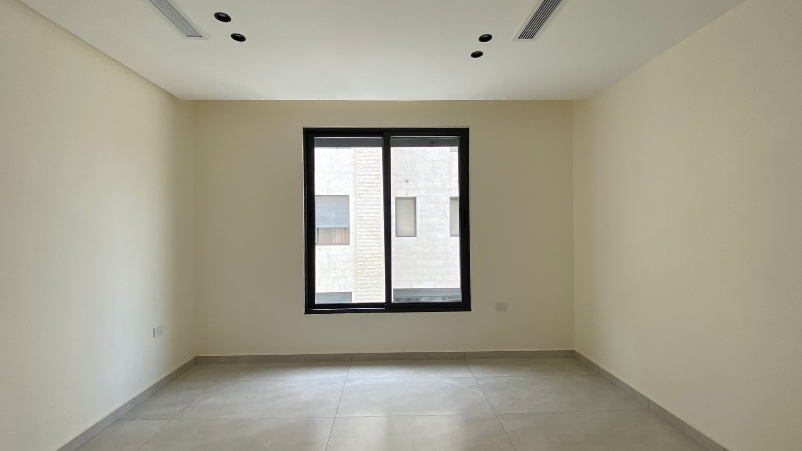 fa1cda9a-8d5b-4089-b2f8-19352d0cd239 1 Empty beige room with a large central window, tiled light floor, and a ceiling featuring recessed black circular fixtures. Window reveals a nearby building exterior.