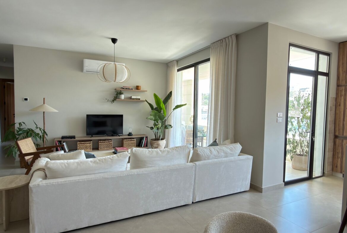 Bright, open-concept living room with a white sofa facing a TV stand and shelves, greenery, and large glass doors to a balcony on the right side