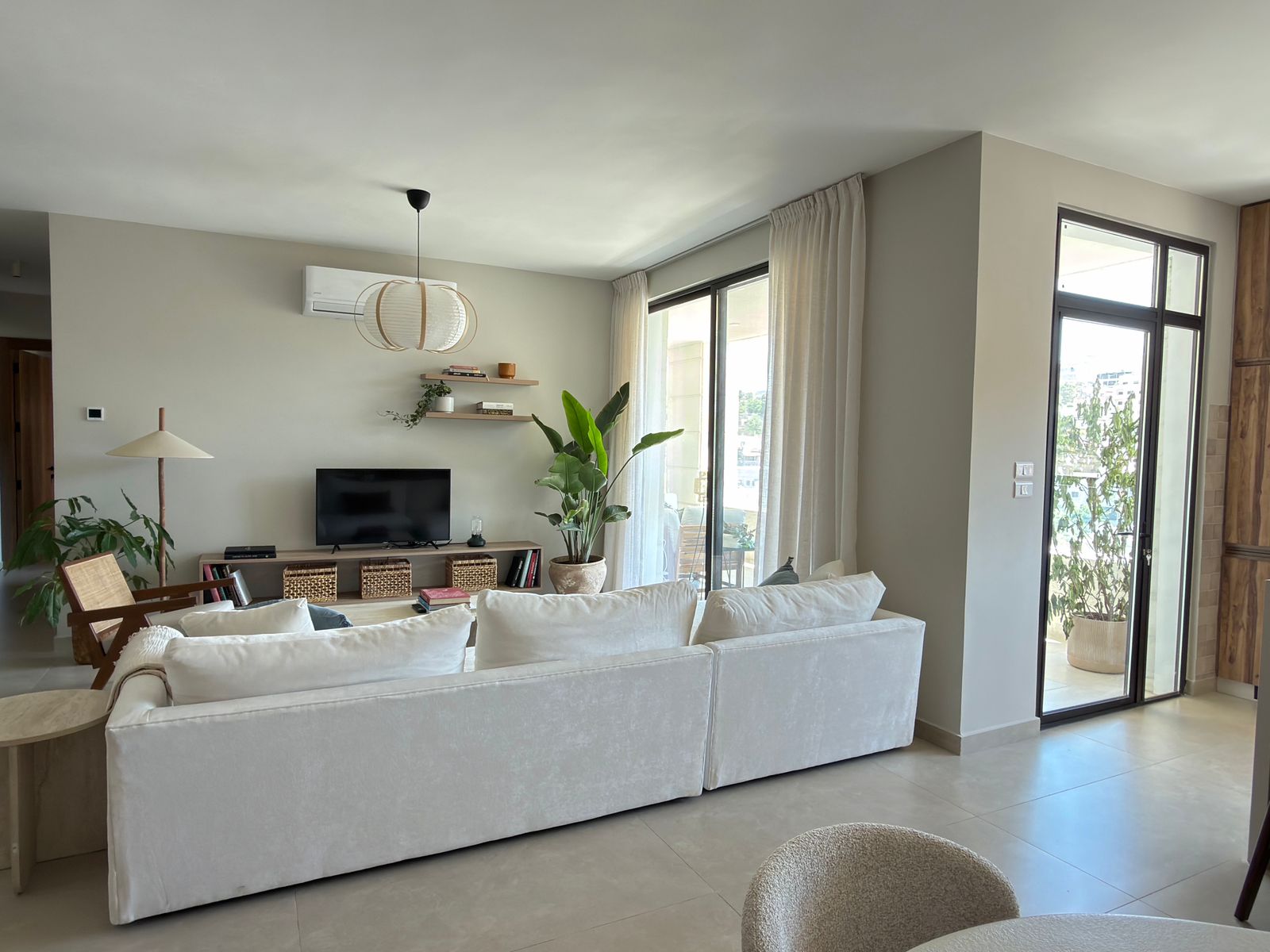 fa8ebc3d-e403-496d-be32-31ce90b7f572 Bright, open-concept living room with a white sofa facing a TV stand and shelves, greenery, and large glass doors to a balcony on the right side