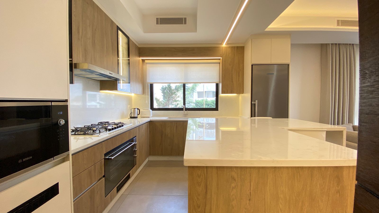 fb724a11-e12e-4b16-88f7-801673a61fc4 Modern kitchen with white quartz island, wood cabinetry, stainless steel fridge, gas cooktop and built-in oven.