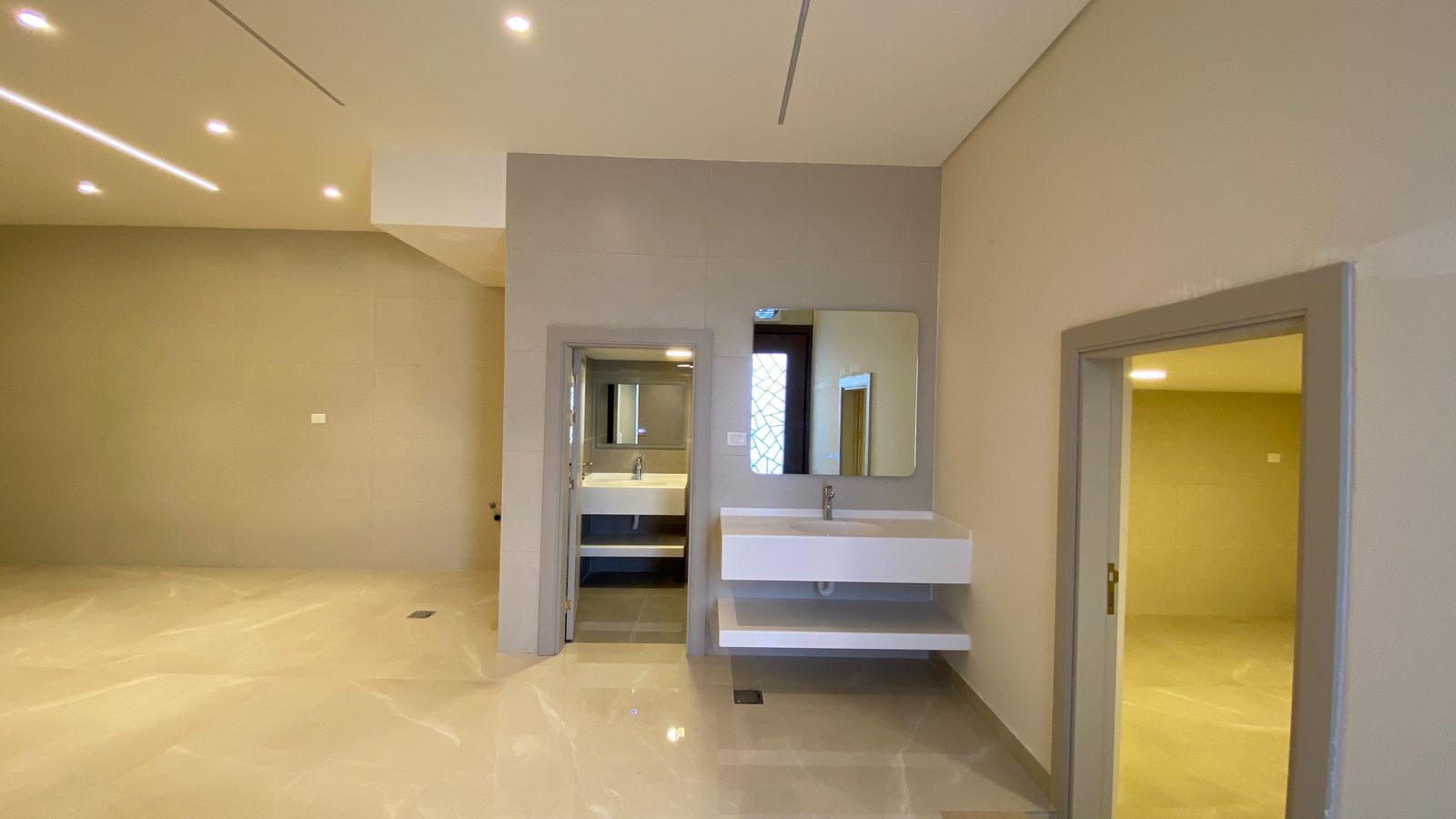 fc46fa67-f660-4fa6-ab30-361fd9c38ffc A modern restroom area with a floating white vanity and rectangular mirror, beige walls, and glossy tile floors; doorways lead to adjoining rooms.