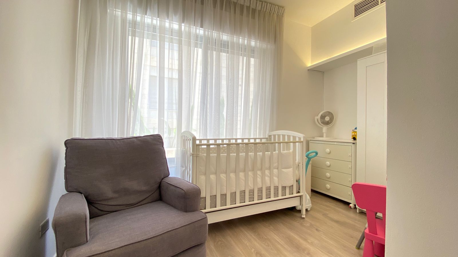 fcf5eb67-80d6-4234-92d8-745bbac0e6e7 Bright nursery with white crib, grey armchair, and a white dresser by a window with sheer curtains and a small pink chair nearby.