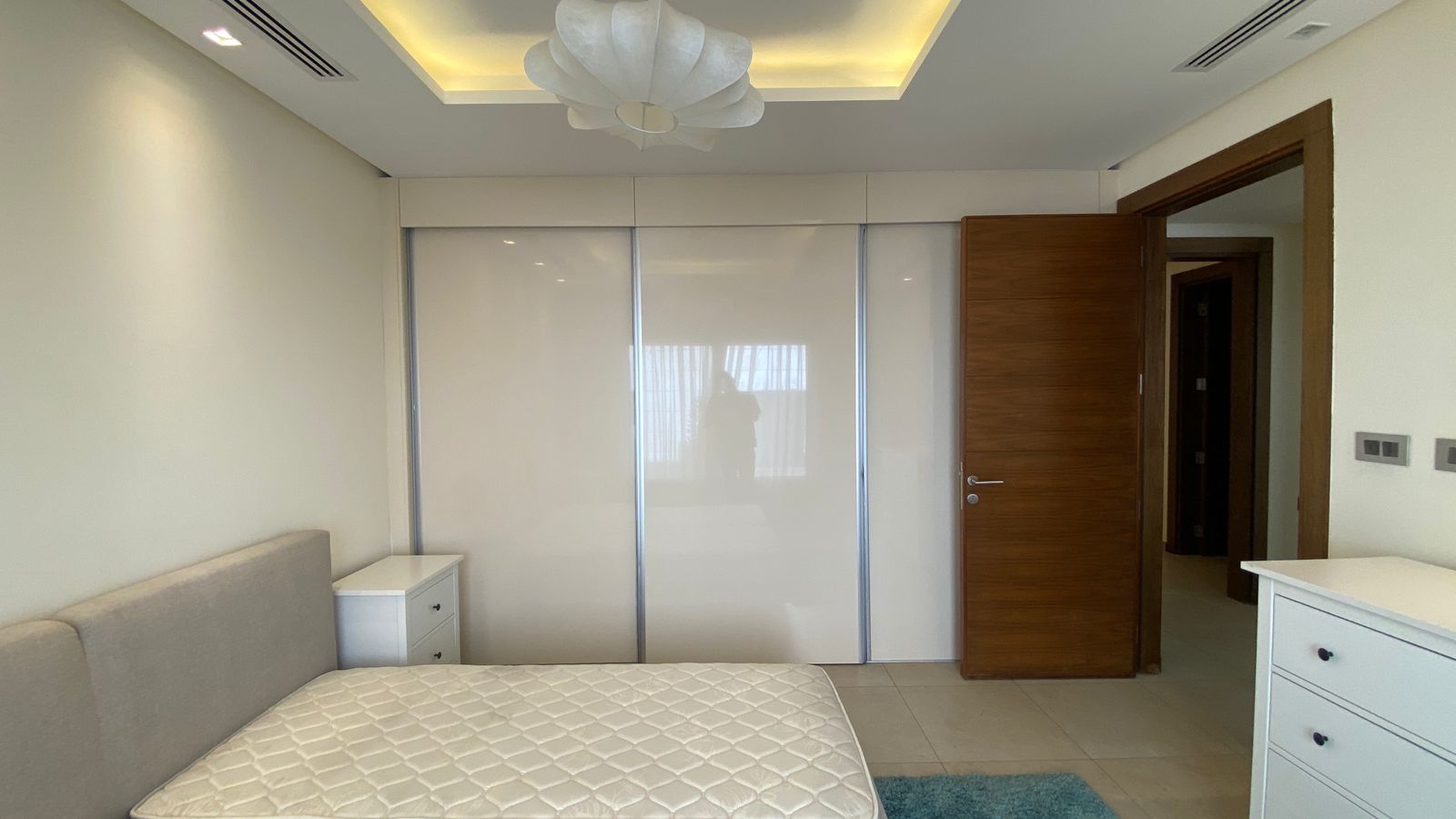 fd3aed21-b7f2-4f2b-8afb-a6151146df3a Modern minimalist bedroom with white sliding wardrobes, a beige upholstered bed, a white nightstand, and an open wooden door.",