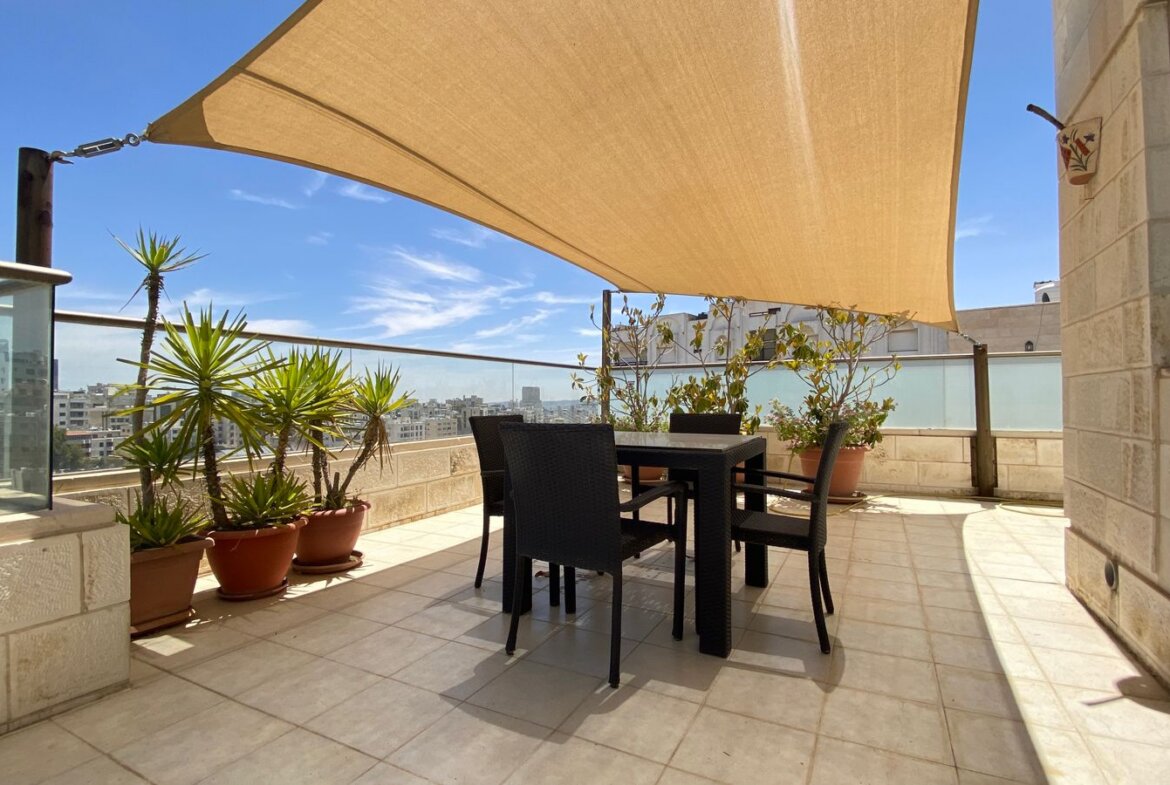 Rooftop terrace with a beige fabric shade sail, black dining table and chairs, and potted plants against a city skyline view.