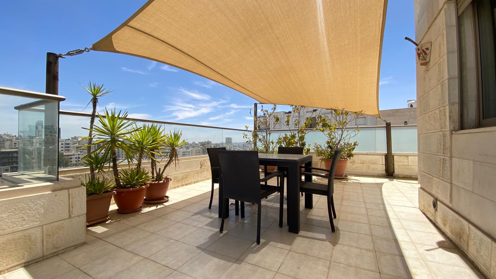 fd4e6131-b97b-4010-89dd-e825d1c9927e Rooftop terrace with a beige fabric shade sail, black dining table and chairs, and potted plants against a city skyline view.
