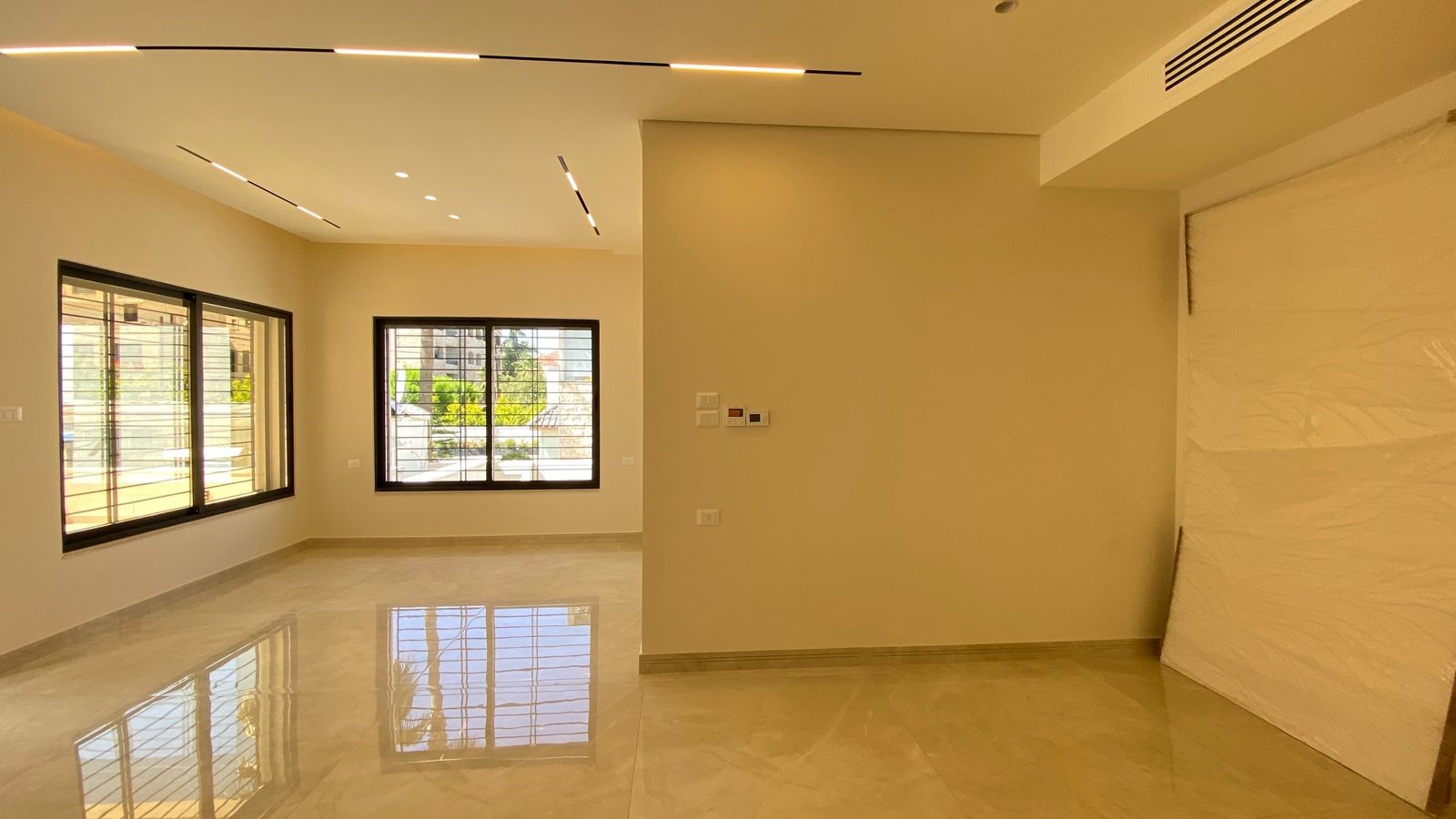 fd9e8341-ae60-480a-888e-a6c9f1231021 Empty modern living room with beige walls, large windows, and a glossy tile floor reflecting light from ceiling fixtures.