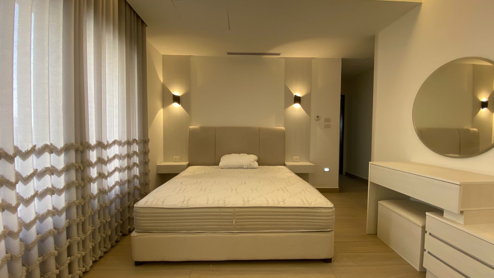fda2cbb4-0ea6-4f5e-b424-3e9ee148739f Modern beige bedroom with a king-size bed, padded headboard, and sheer curtains along the left wall.