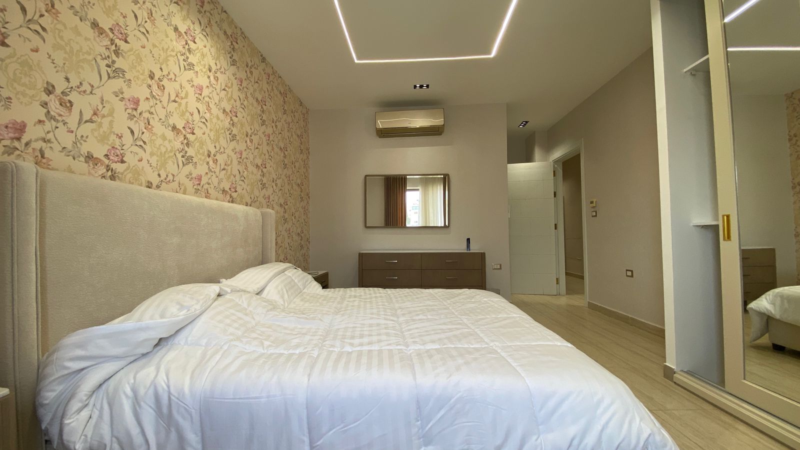 fdce9805-1d16-4de8-b854-bec4b939ce8c Bedroom with a floral wallpaper wall, large upholstered bed, and white duvet; dresser and mirror opposite; modern ceiling light fixture and wall air conditioner visible.