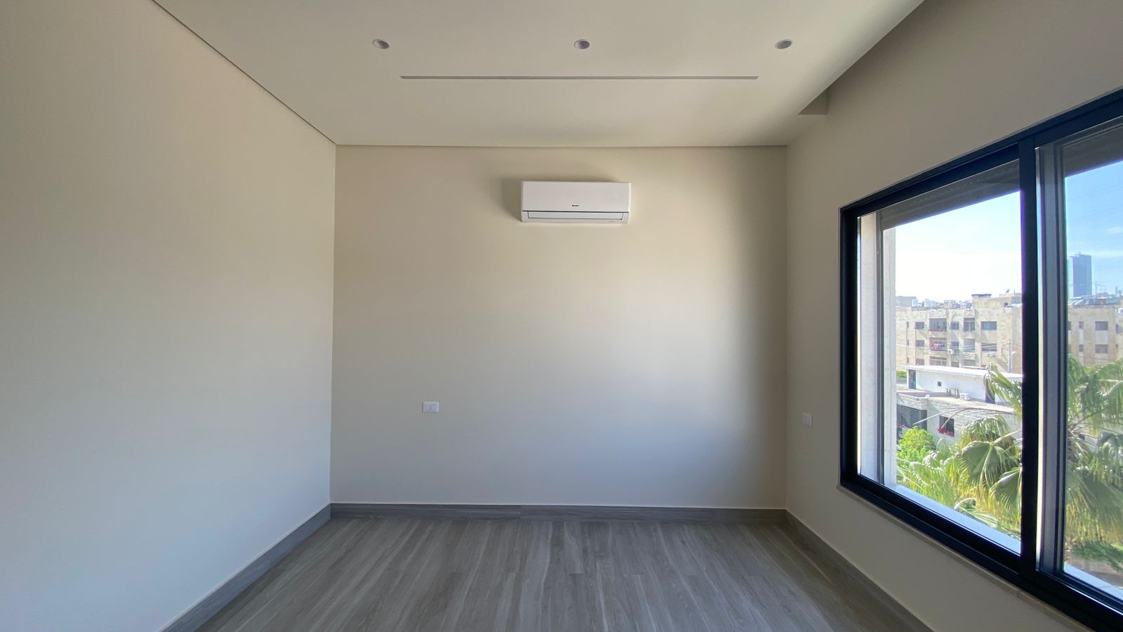 fe8b605e-60e7-451c-8296-474464a3e5d6 Empty modern room with gray walls, wood-like flooring, a wall-mounted air conditioner, and a large window showing a cityscape with palm trees outside.