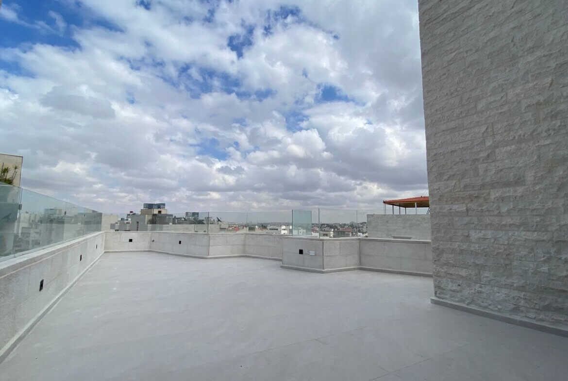 Rooftop terrace with glass railings, pale stone walls, and a cloudy blue sky over a distant city skyline.
