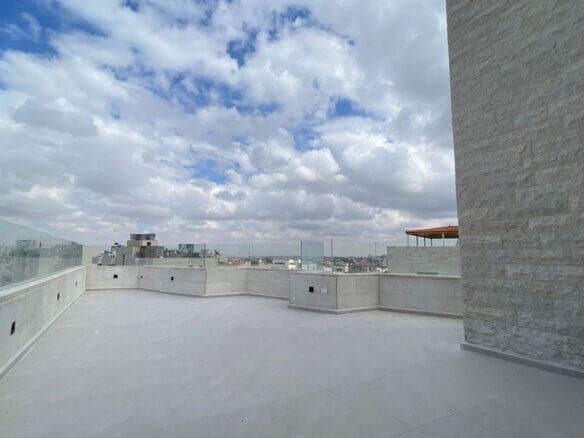 Rooftop terrace with glass railings, pale stone walls, and a cloudy blue sky over a distant city skyline.