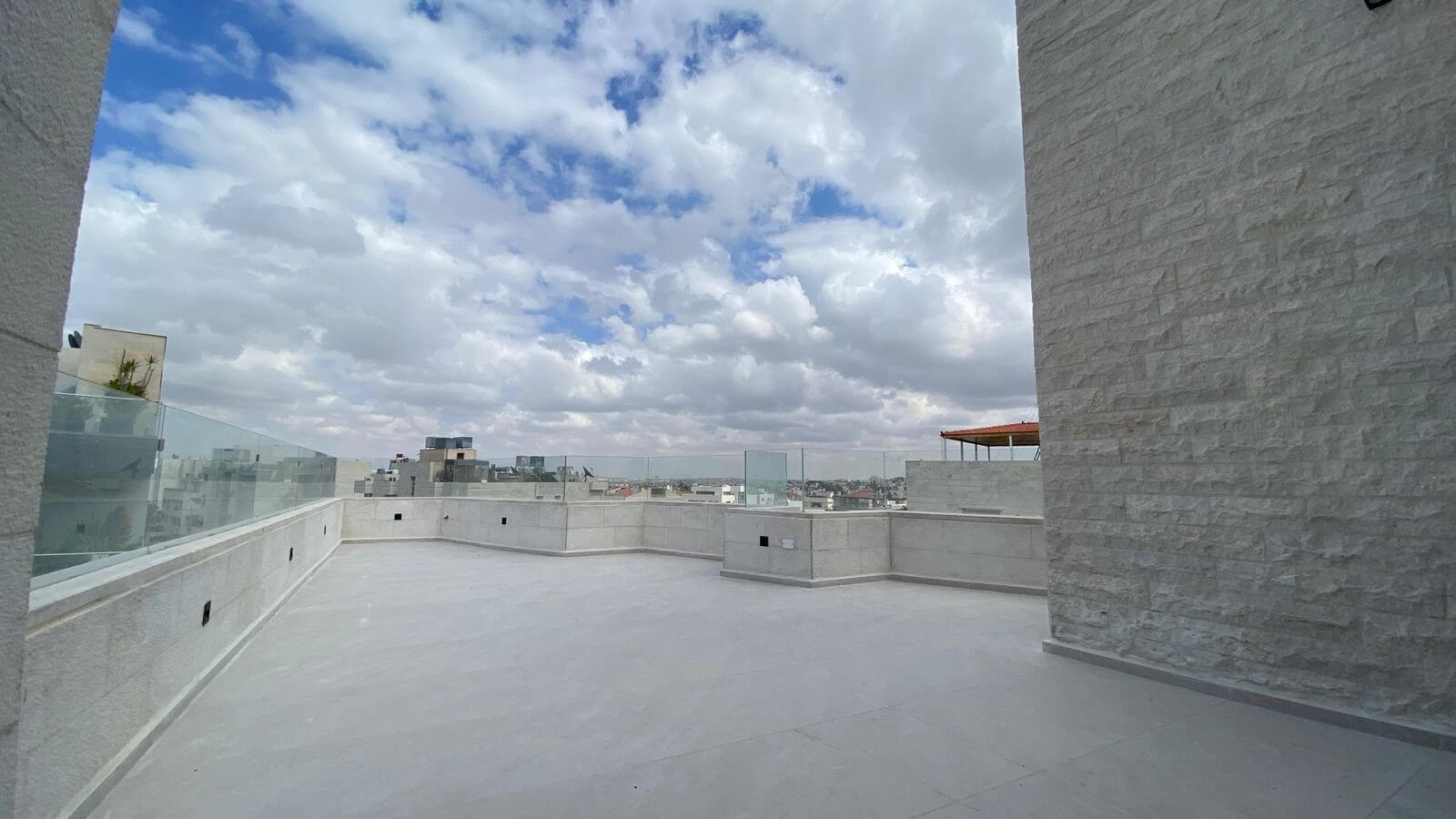 Rooftop terrace with glass railings, pale stone walls, and a cloudy blue sky over a distant city skyline.