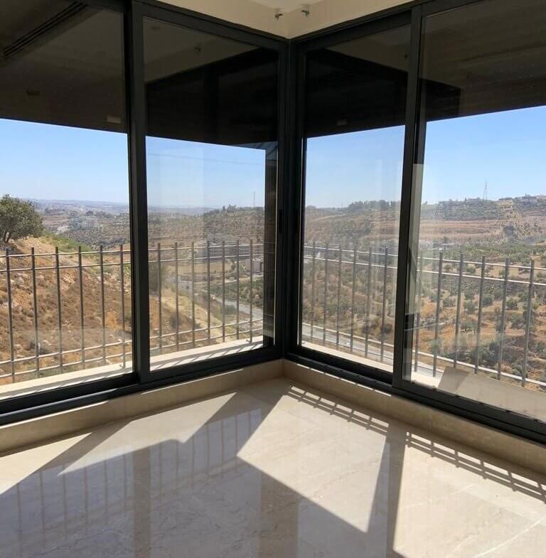 Corner sunroom with black-framed glass walls and a railing, overlooking a hilly landscape; sunlight casts geometric shadows on the polished floor.