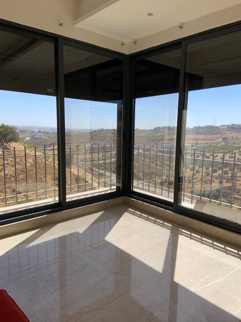 ffd965df-fbd6-4066-894a-30aa97ff200f Corner sunroom with black-framed glass walls and a railing, overlooking a hilly landscape; sunlight casts geometric shadows on the polished floor.