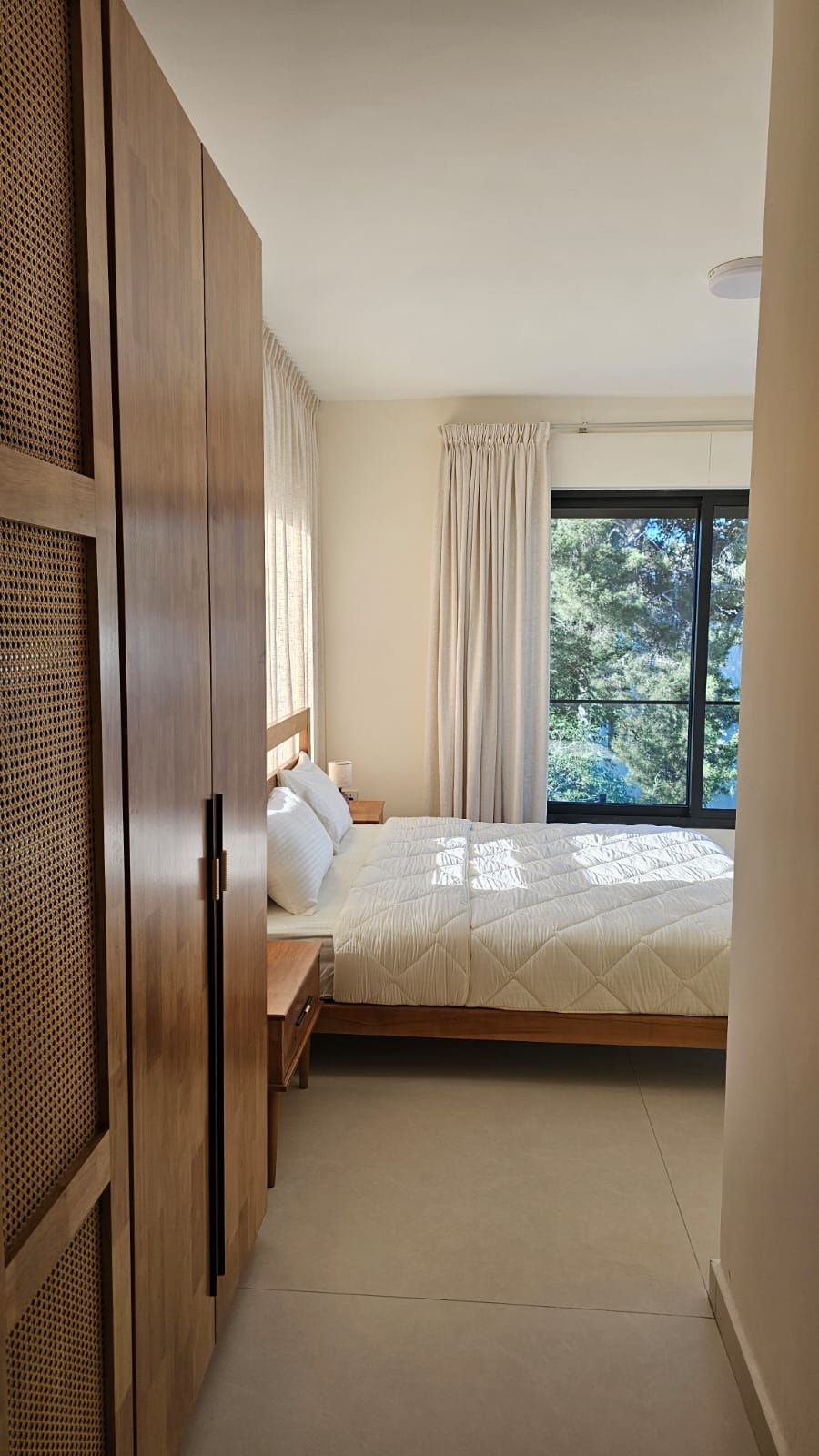 0331972a-5bff-412d-8544-af13219e2239 Sunlit bedroom with a tall wooden wardrobe along the left, a white quilted bed, and a window with beige curtains.