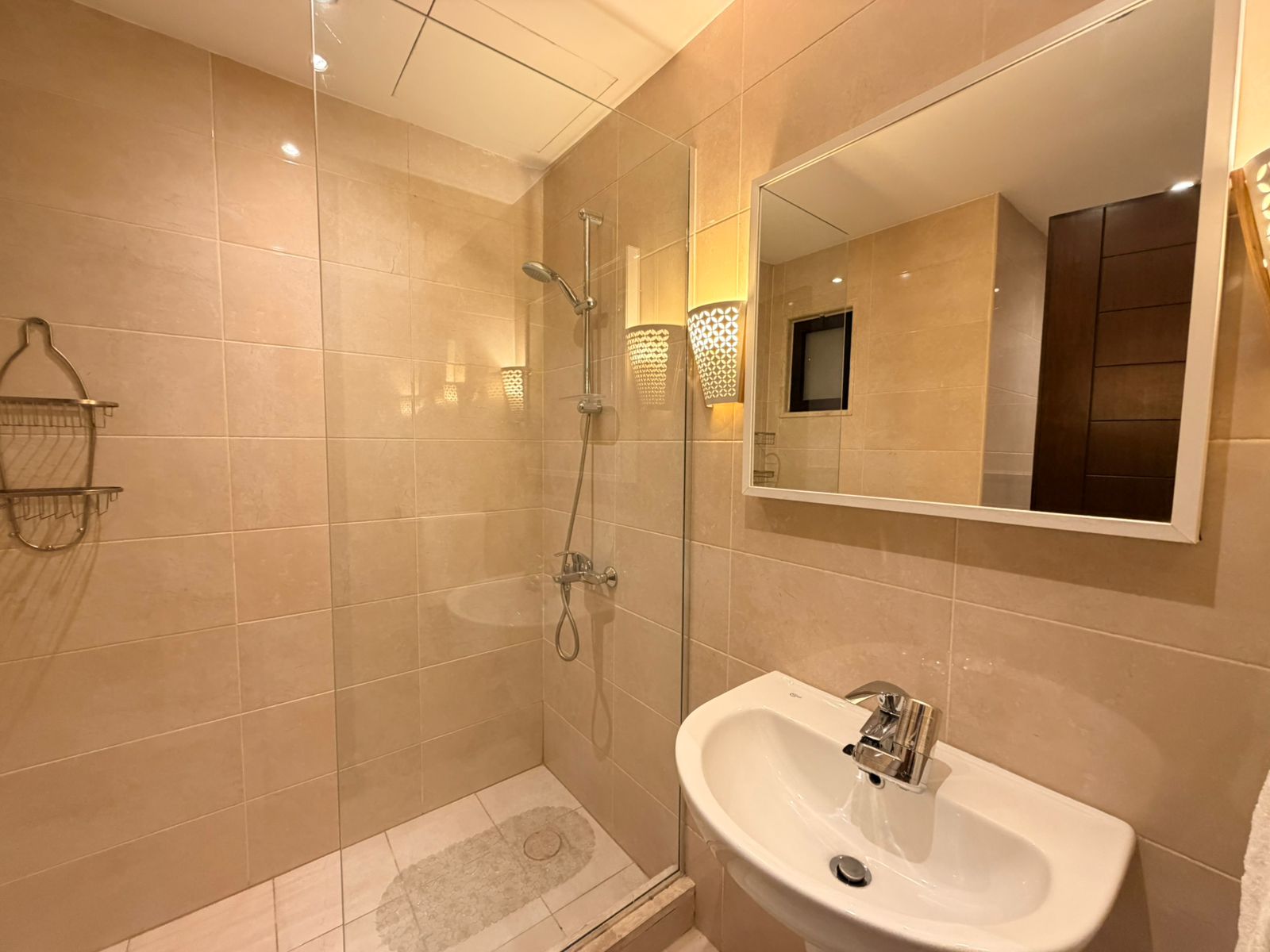 036739b0-44a6-4253-b256-5bef9c8b34a5 Beige-tiled bathroom with a glass shower enclosure, wall mirror, and white sink.