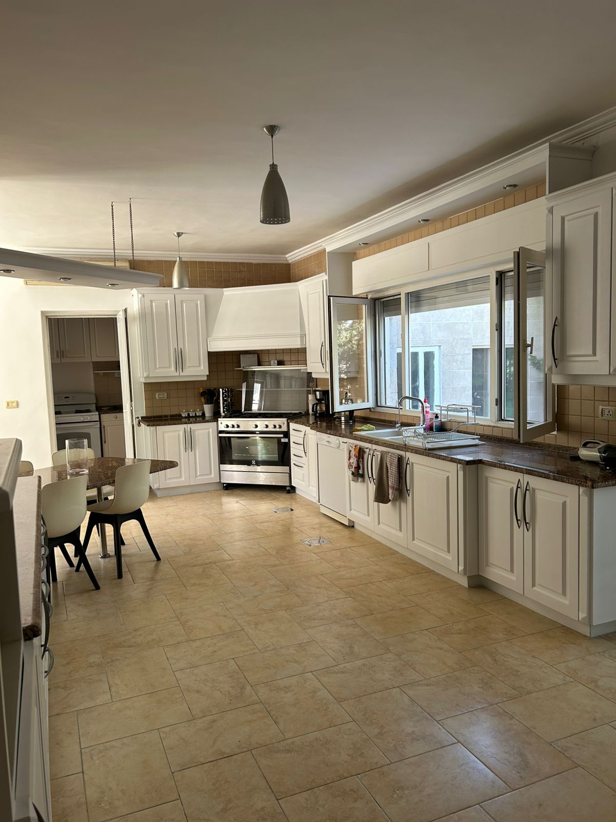 03a8b8a2-af93-48a4-b693-ba3c6d1d27e8 Bright L-shaped kitchen with white cabinets, beige tile floor, and a stainless steel stove under a range hood with a window above the sink.