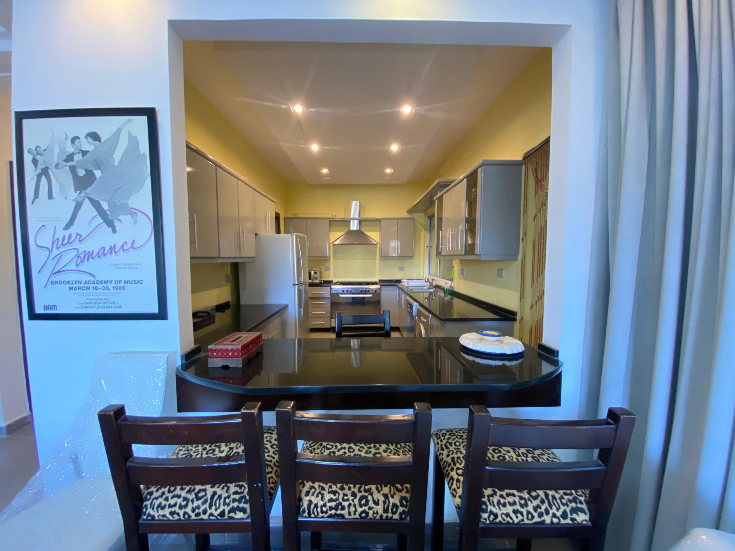 0431a218-d112-4a4e-92c4-1aa5b1f9ecda Open kitchen with yellow walls, stainless appliances, and leopard-print bar stools along a dark counter viewed through an opening from a dining area.