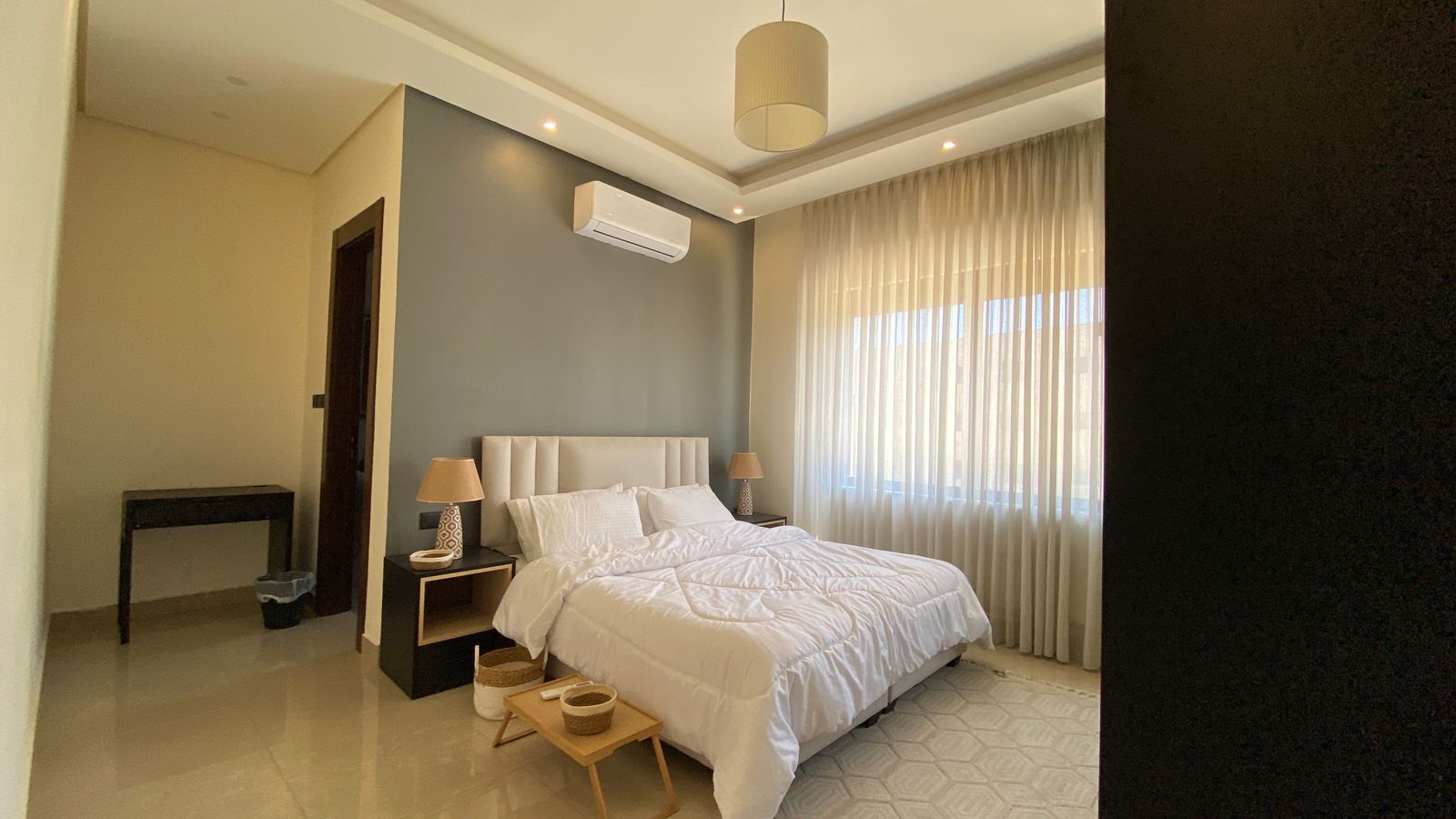 064f1059-5af9-4552-b8ac-ada76d6e42cf Bright hotel bedroom with a white bed, tufted headboard, two matching nightstands and lamps, and sheer-curtained window letting in soft light.