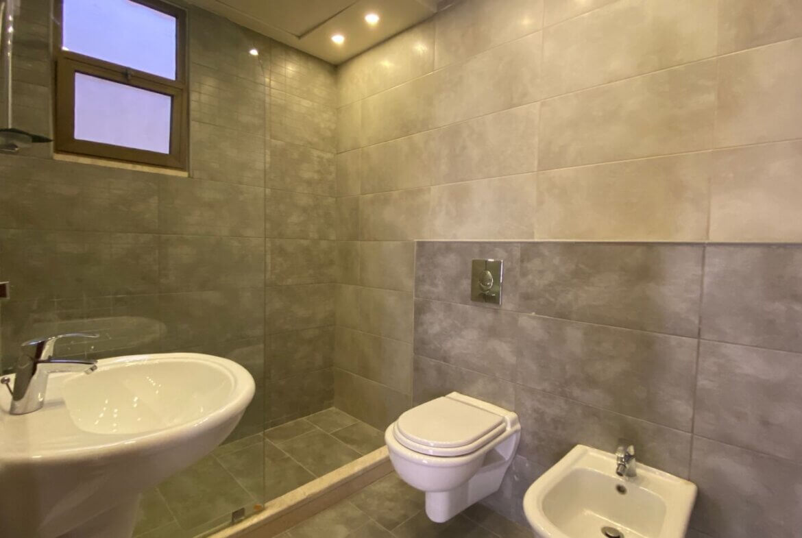 0663a2ac-95c7-4b77-9ce6-fa8f00884787 Modern bathroom with a wall-mounted toilet, a bidet, and a curved white sink beside a glass shower enclosure in gray tiles.