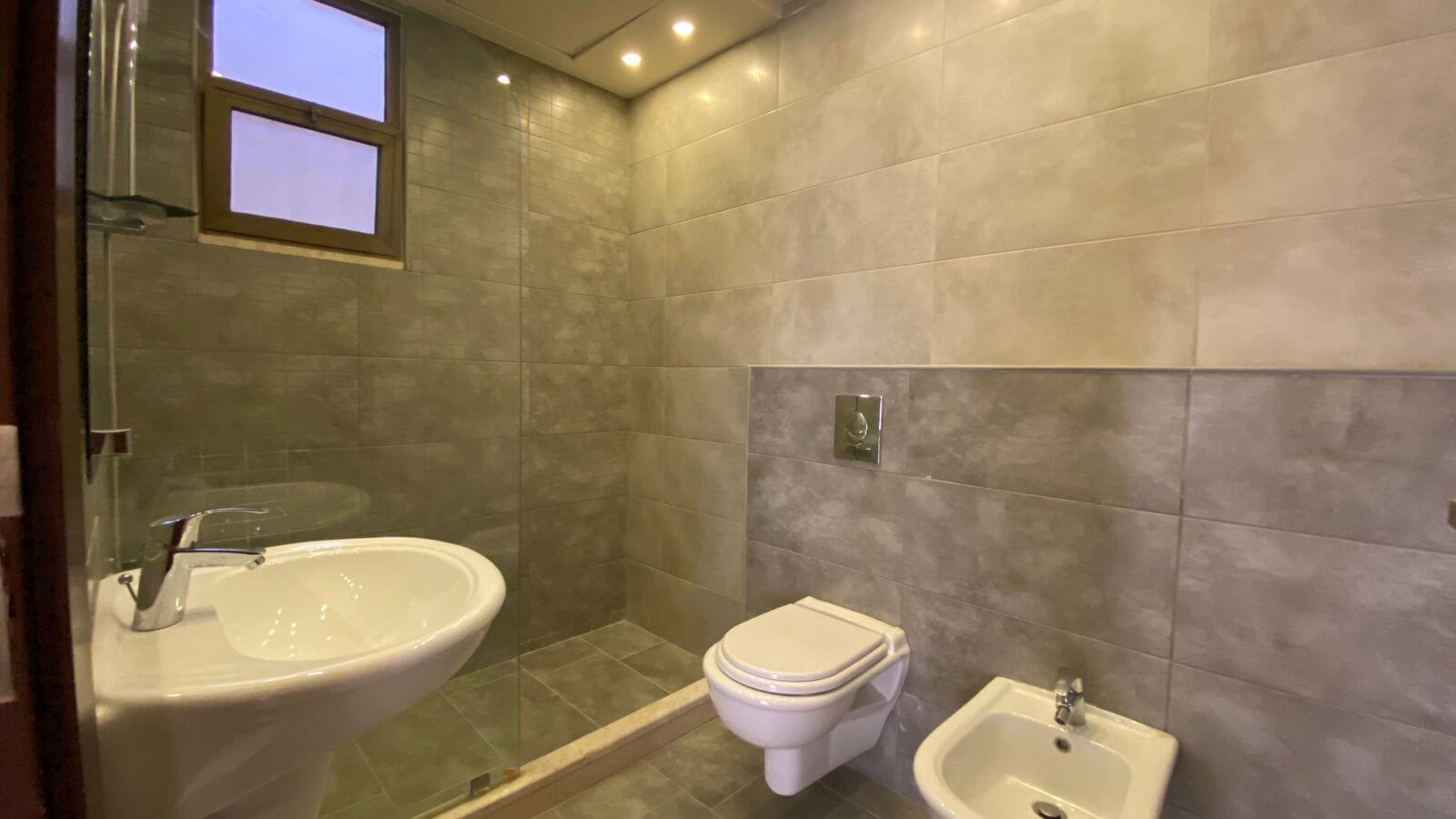 0663a2ac-95c7-4b77-9ce6-fa8f00884787 Modern bathroom with a wall-mounted toilet, a bidet, and a curved white sink beside a glass shower enclosure in gray tiles.