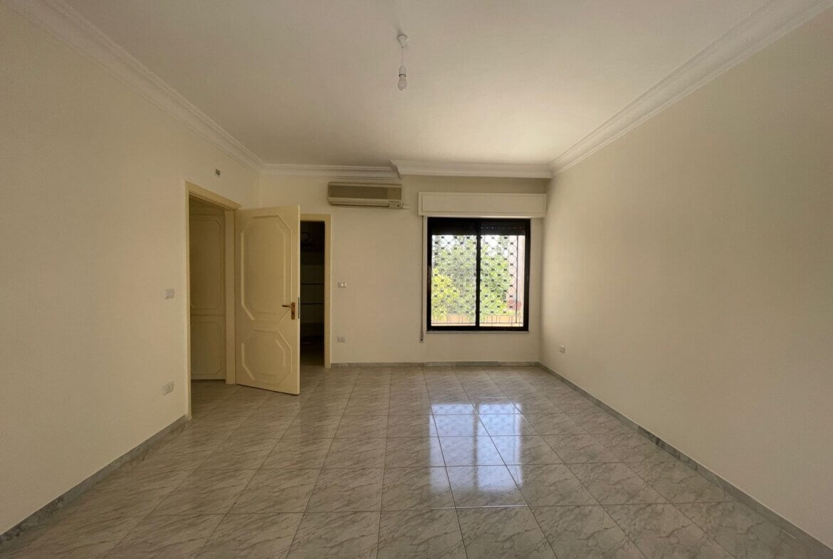 079b03fe-b1a3-4c7b-89d2-6ba7dda90b99 Bright, empty living room with white walls, gray marble-tiled floor, an open door to the left, and a window with a metal grid on the far wall.