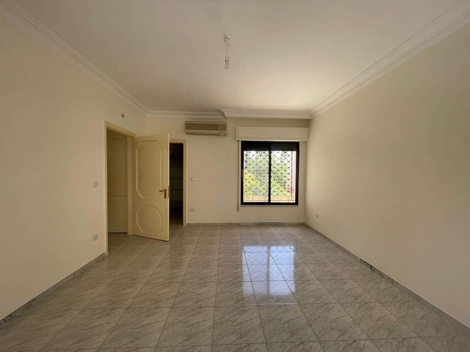 079b03fe-b1a3-4c7b-89d2-6ba7dda90b99 Bright, empty living room with white walls, gray marble-tiled floor, an open door to the left, and a window with a metal grid on the far wall.