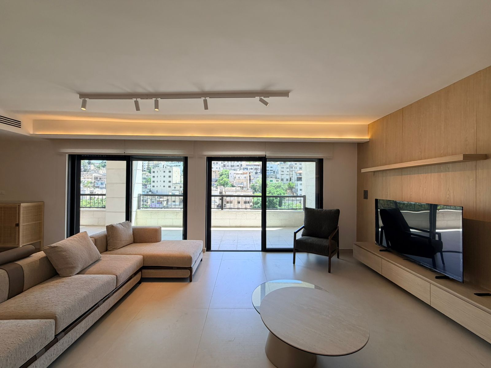 07c4dfa8-17b1-4c10-b70f-4830a28a9729 Modern living room with a beige sectional sofa facing a large windowed balcony and a flat-screen TV on a wood-paneled wall. Nearby chair and glass coffee table create a minimalist setup.