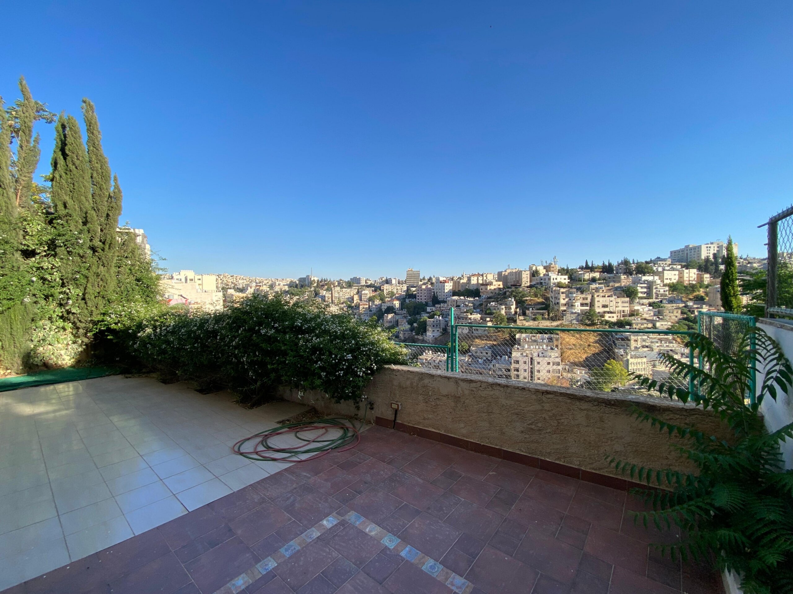 07cb7ad1-81bb-4df4-8279-0767ae2fc9bc Terrace with tiled floor and a cityscape beyond, tall trees on the left under a clear blue sky.