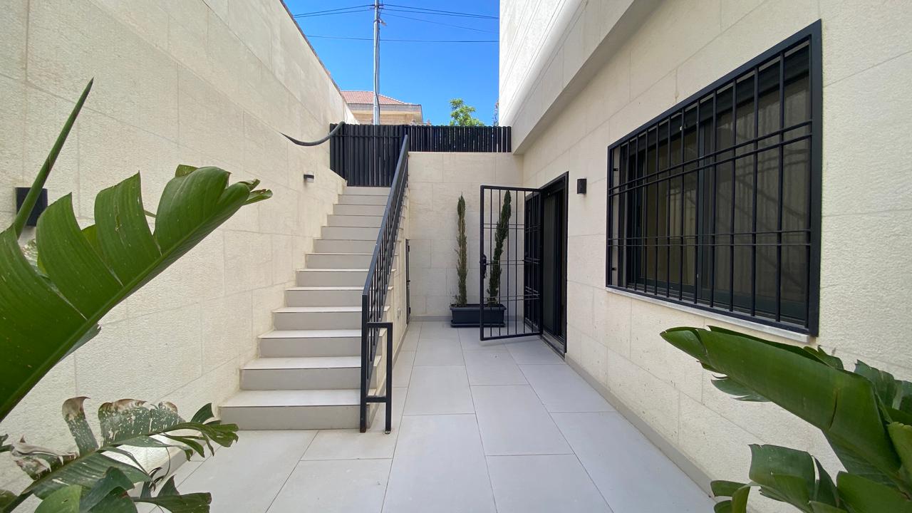 07cf9b66-d1cc-415b-80f7-98d4c391a2a6 Courtyard with beige stone walls, a left-side staircase and black railing, a gated doorway, and tall potted plants near the entrance grid window.