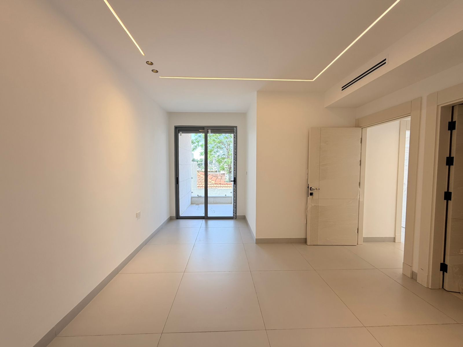 084215f3-2ecc-4b68-b0e1-63d82e9f67c5 Empty, modern, unfurnished room with a sliding glass door to a balcony and visible trees outside; white walls and tiled floor with recessed ceiling lights present.