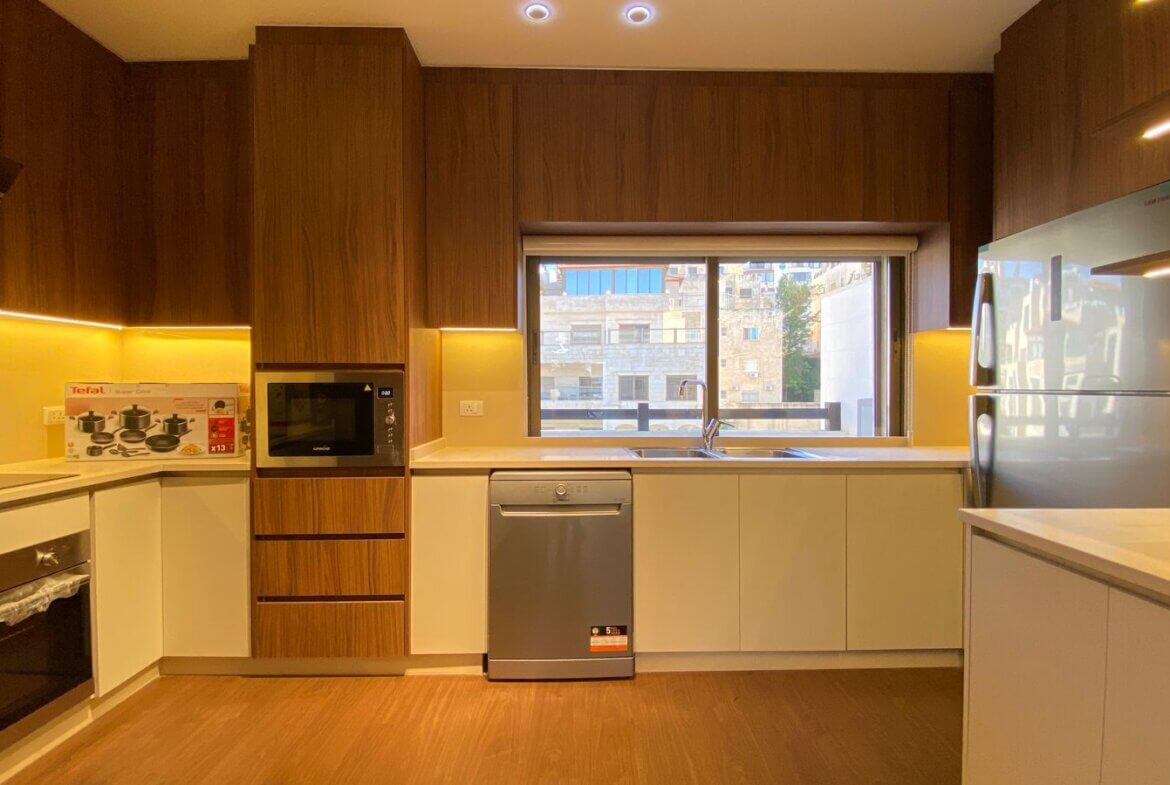 Modern kitchen with dark wood cabinets, white lower cabinets, and warm under-cabinet lighting; built-in microwave, stainless dishwasher, oven, and side-by-side fridge visible by a large window overlooking buildings outside.