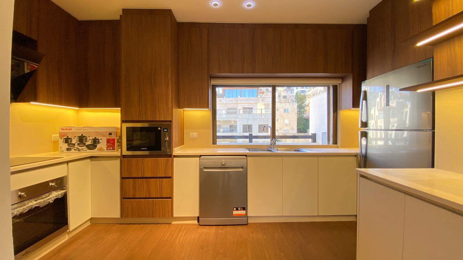 Modern kitchen with dark wood cabinets, white lower cabinets, and warm under-cabinet lighting; built-in microwave, stainless dishwasher, oven, and side-by-side fridge visible by a large window overlooking buildings outside.