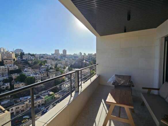Balcony with a wooden table and cushioned seating, overlooking a dense cityscape of white buildings on a sunny day.