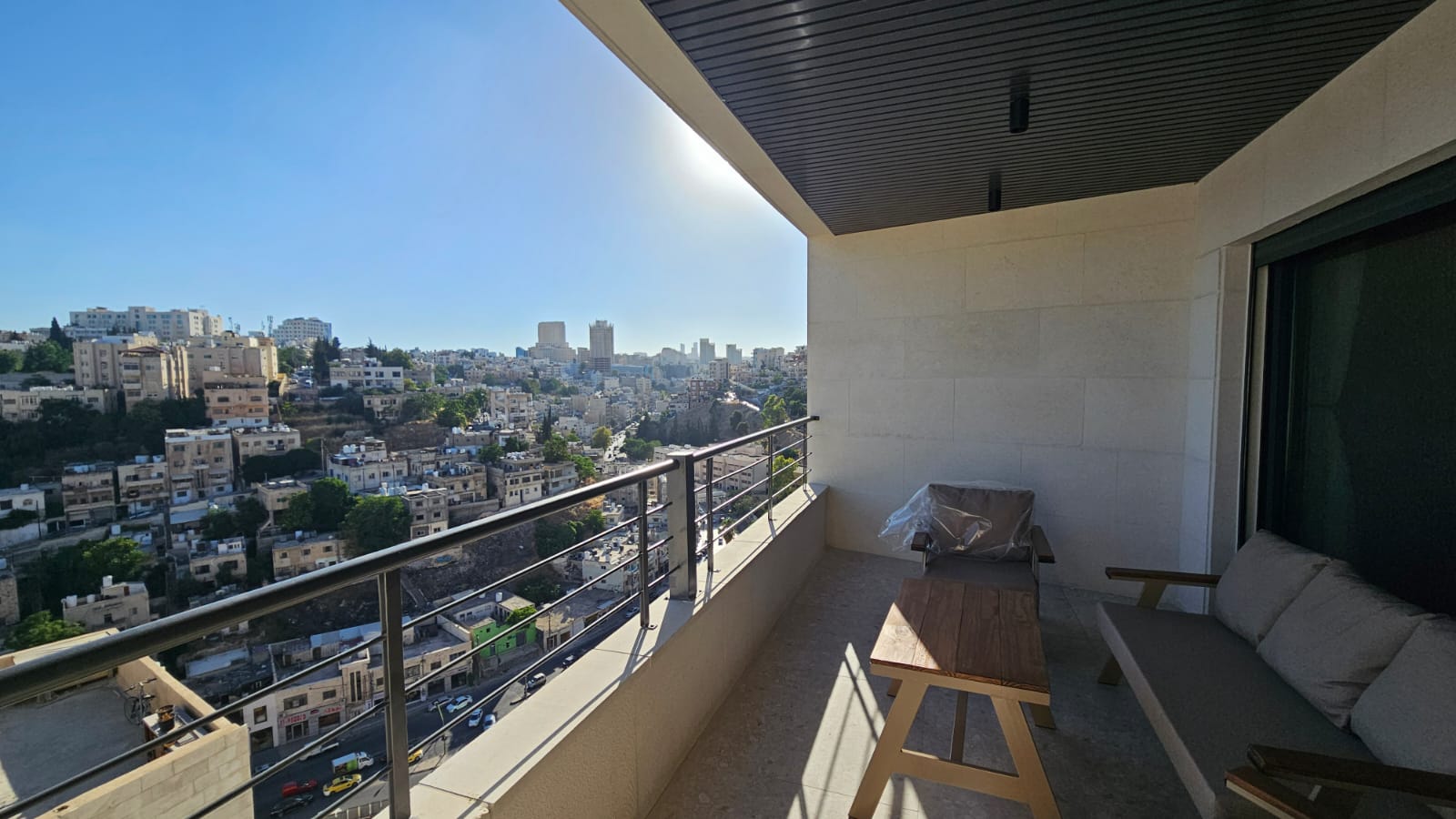 09386136-2cfe-4ed6-a24e-3732dfb3d94a Balcony with a wooden table and cushioned seating, overlooking a dense cityscape of white buildings on a sunny day.