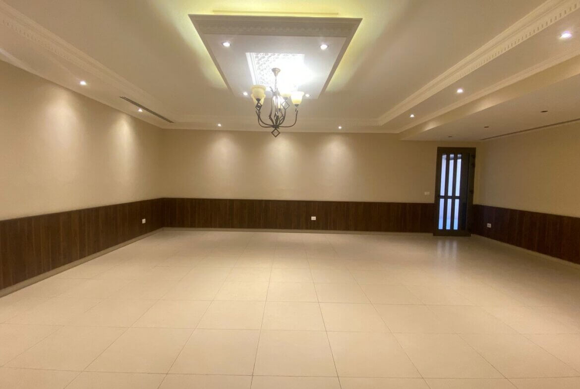 0961a3b0-1db2-43cf-ae26-748fca7c6cab A large empty living room with beige walls, dark wood wainscoting, tiled floor, and a chandelier hanging from a decorative ceiling panel. A glass-paneled door is on the right side.