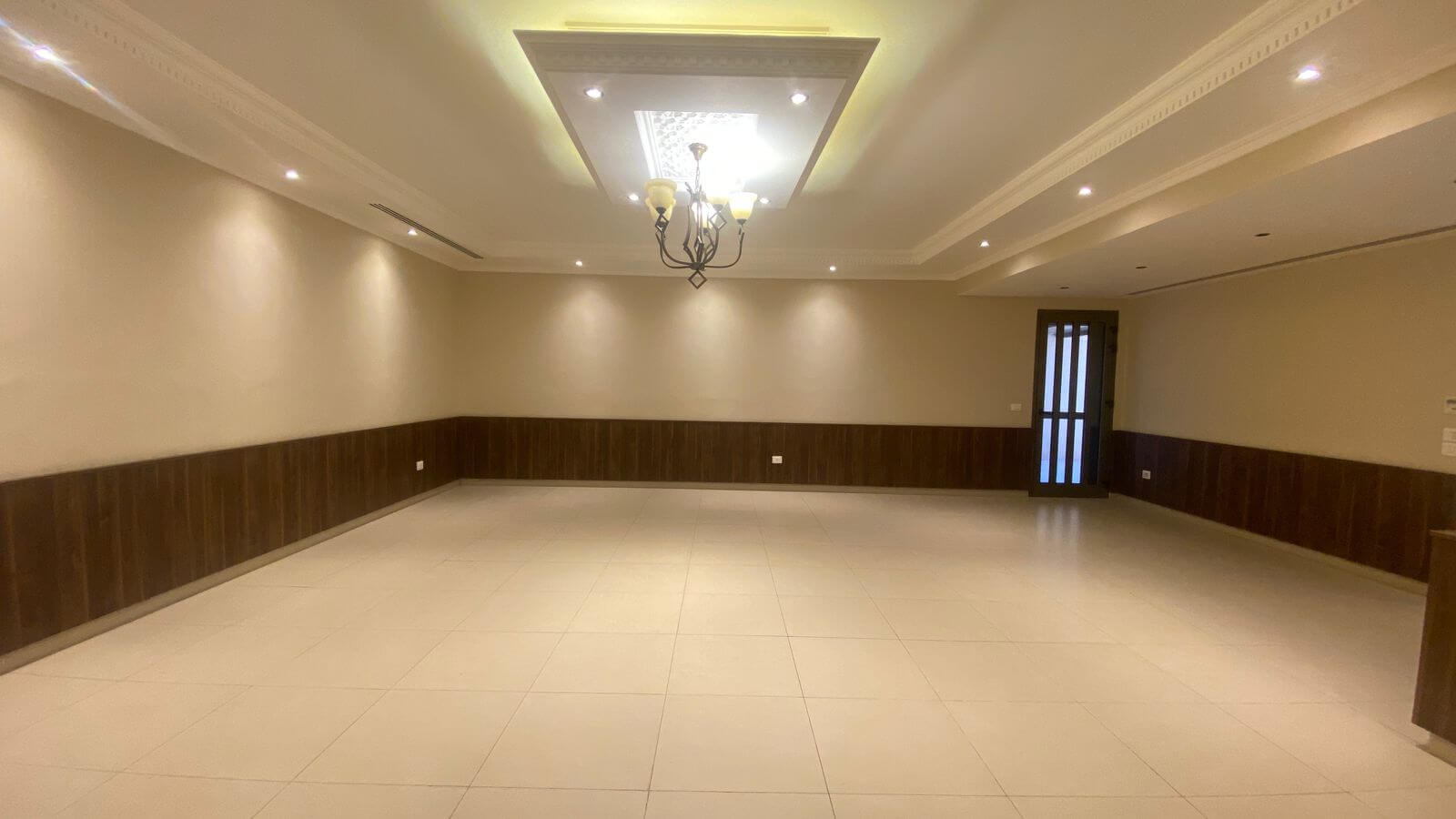 0961a3b0-1db2-43cf-ae26-748fca7c6cab A large empty living room with beige walls, dark wood wainscoting, tiled floor, and a chandelier hanging from a decorative ceiling panel. A glass-paneled door is on the right side.